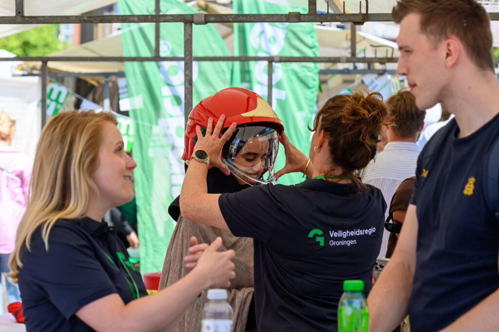 Medewerker van Veiligheidsregio Groningen zet een bezoeker een brandweerhelm op tijdens De Banenmarkt
